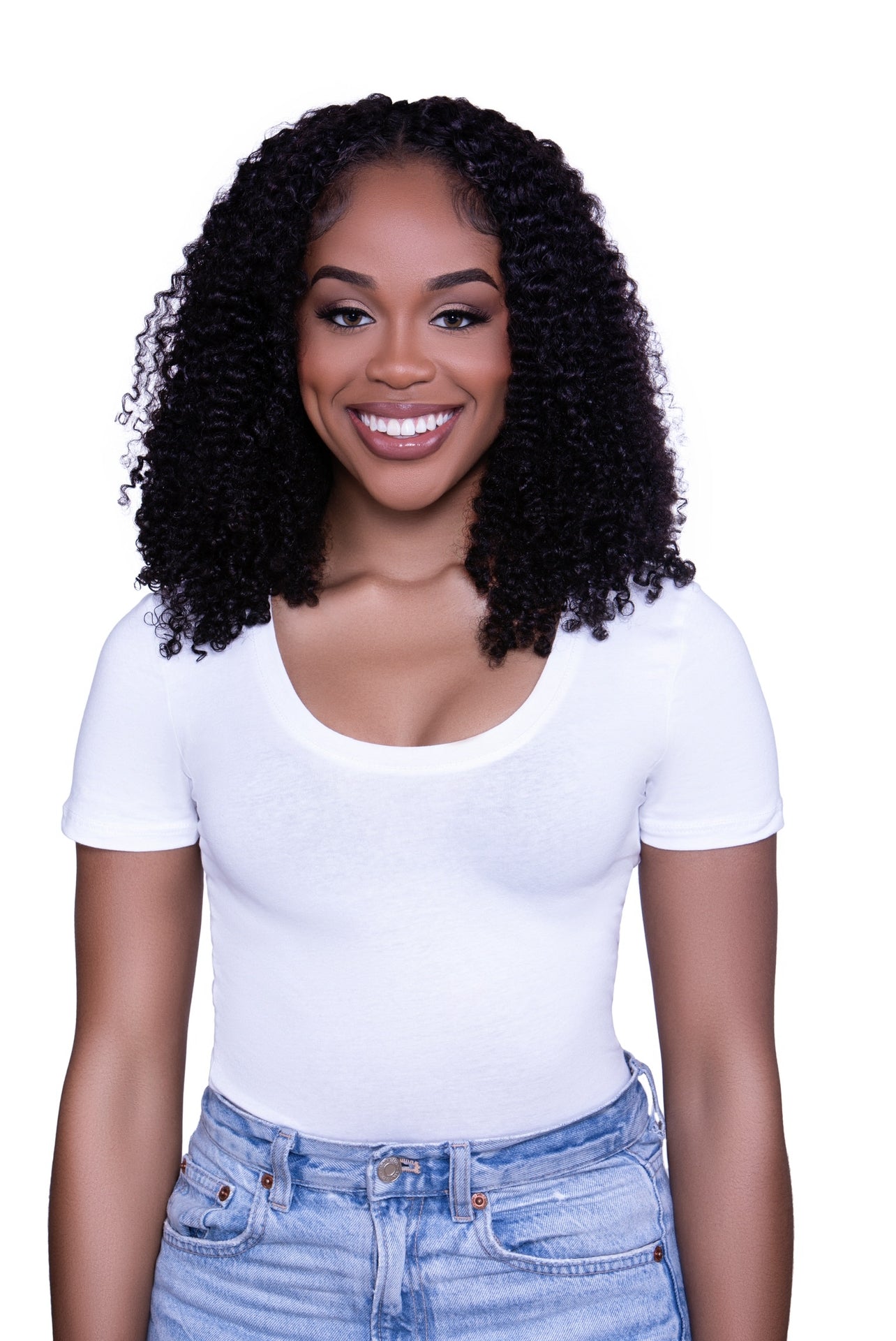 A smiling person with kinky curly hair wearing a white top and blue jeans. They have dark skin and their hair extends to their shoulders.