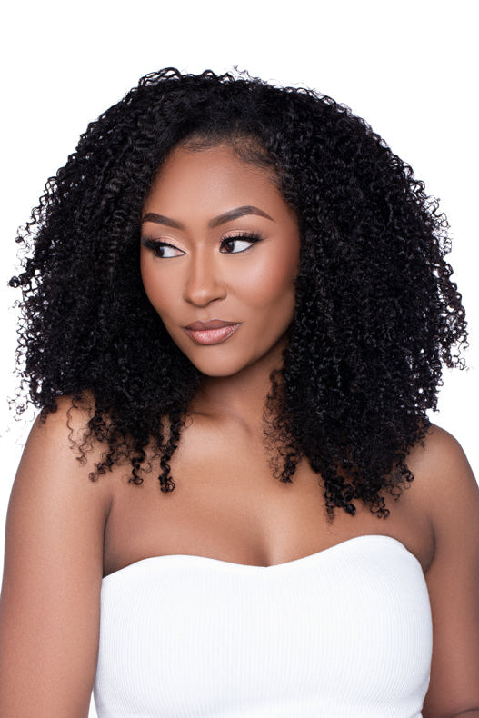 Confident woman wearing the Half Wig ‘Kai’ in a kinky curly texture, styled with voluminous, natural-looking curls and a side part. She poses in a white strapless top, highlighting the full-bodied definition and realistic hairline of the wig.