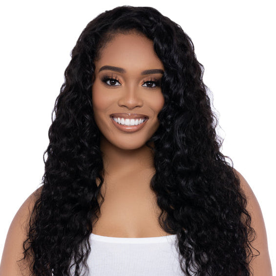 A woman smiling, wearing a white top and black pants, with long loose curly black hair extensions.