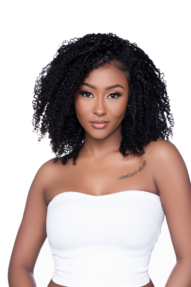 Studio portrait of a model wearing the ‘Kai’ Kinky Curly Half Wig. Her natural curls are blended with the wig's soft, coily texture, creating a seamless transition for a chic, protective hairstyle paired with a simple white bandeau top.