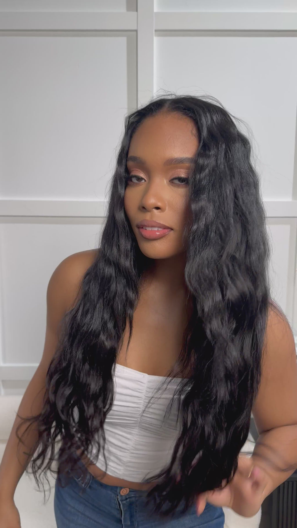 A woman wearing a white crop top smiles as she gently plays with her “Venus” Body Wave Seamless Clip-Ins, beautifully styled with soft, flowing waves that blend effortlessly into her natural hair.