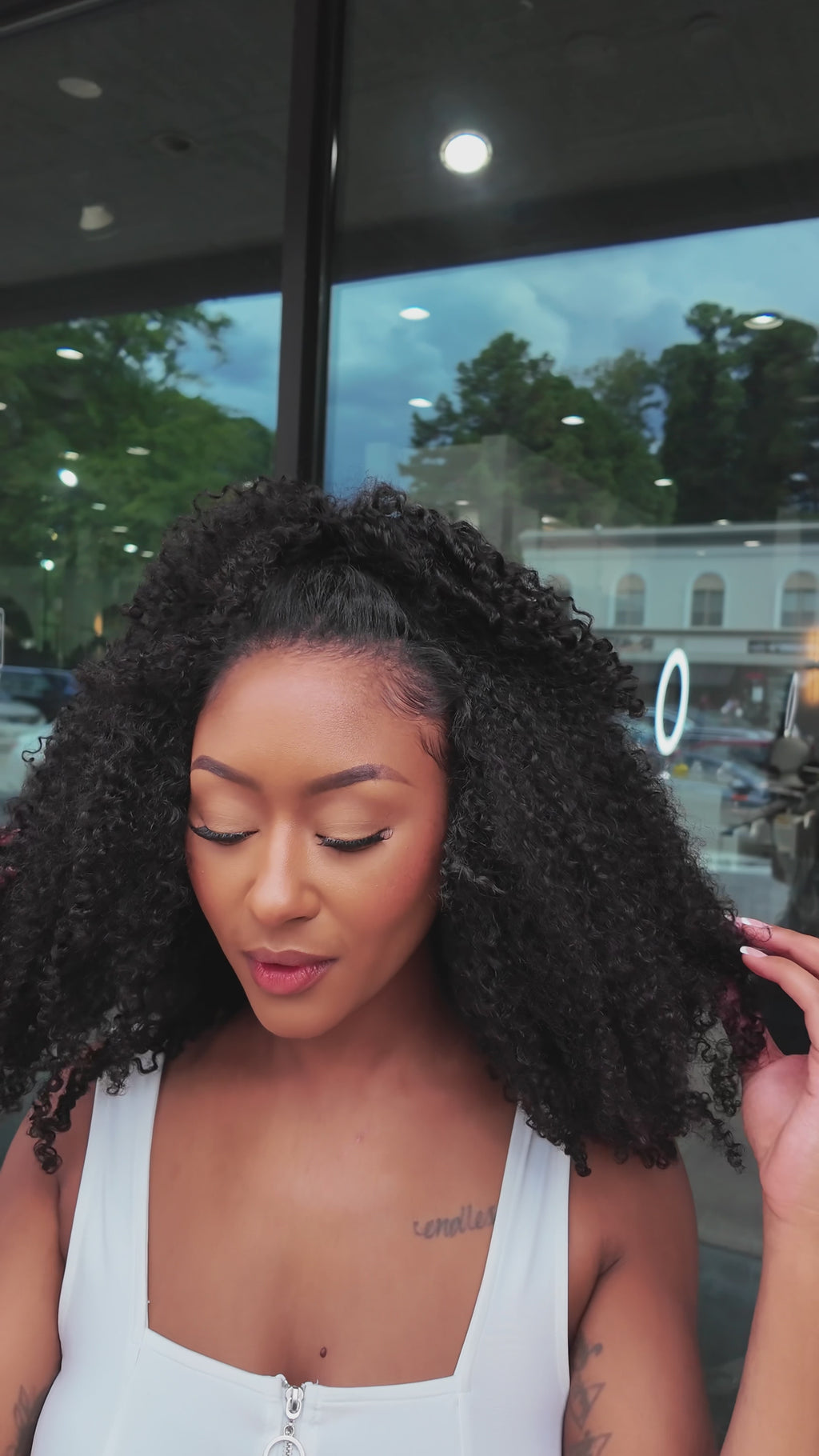 Woman wearing the ‘Kai’ Kinky Curly Half Wig gently fluffing and playing with the voluminous curls, showcasing the soft, bouncy texture and natural hairline.