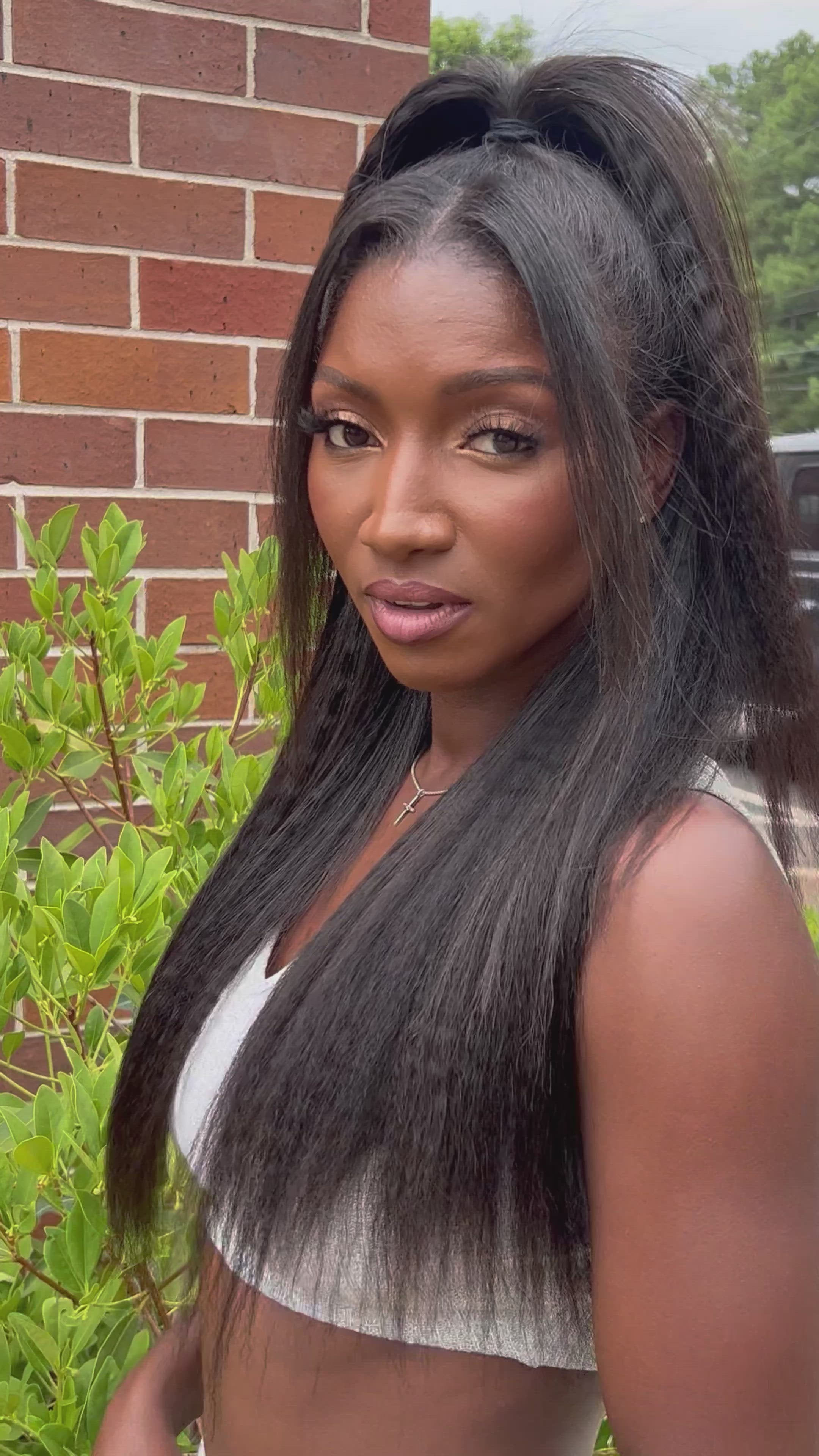 Woman wearing Go Sleek Seamless Kinky Straight Clip-In Extensions styled in a half-up, half-down look. The natural texture blends seamlessly, showcasing volume and length. She's wearing a white tank top with a clean white backdrop.